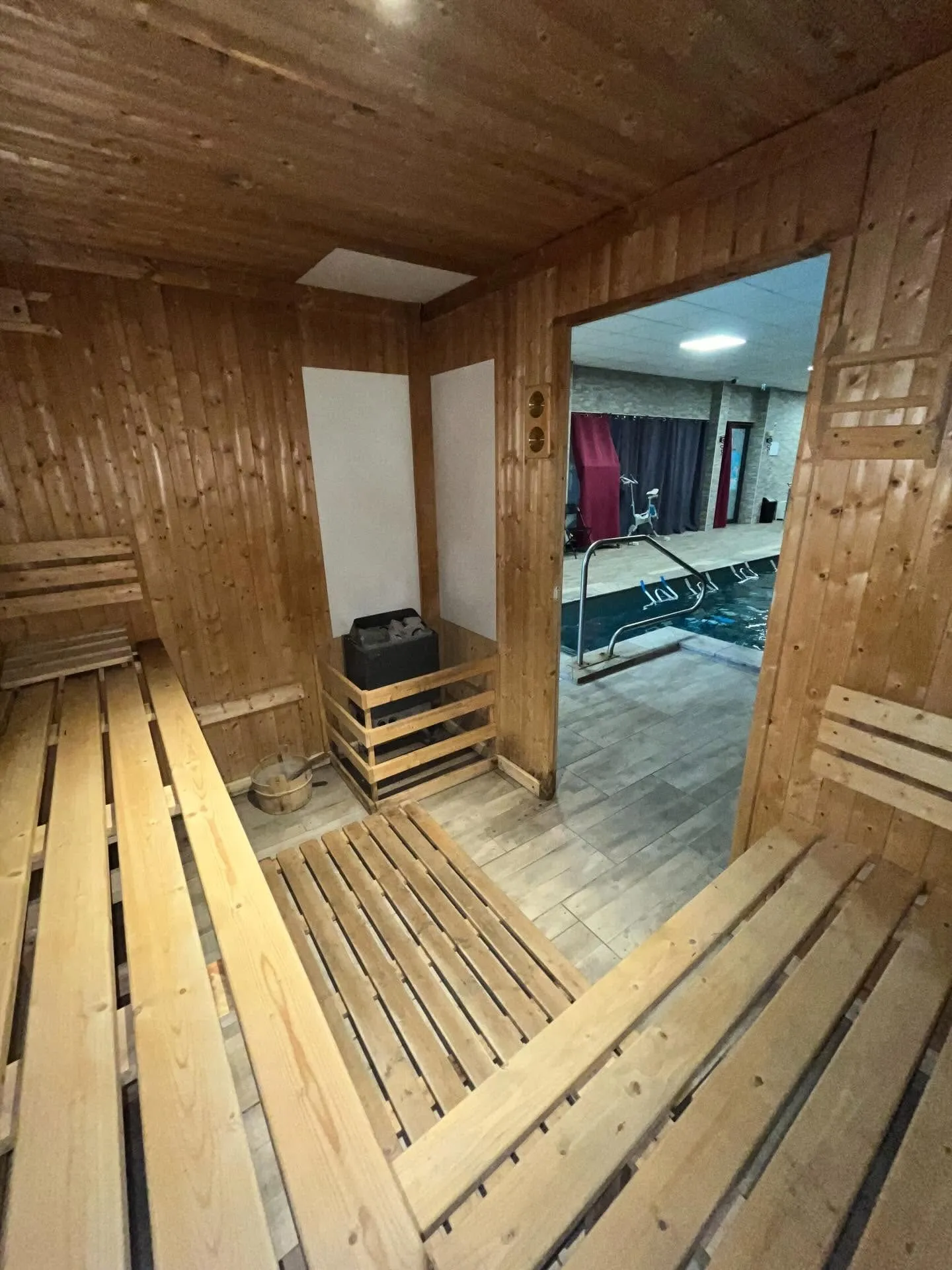Sauna en bois avec ses banquettes et son poêle, porte ouverte sur le bassin